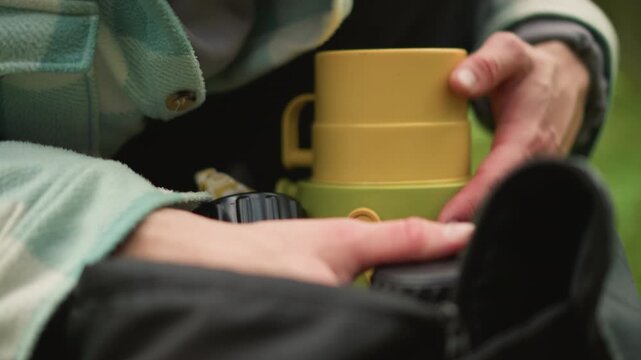 caucasian traveler checking backpack gear before hike, closeup hands adjusting thermos and camera lens, deliberate movements on damp grass, plaid jacket and ring visible, careful inspection
