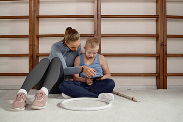 Caucasian woman sitting on floor showing tablet to Caucasian child with Down syndrome, both focused on screen, gymnastic equipment visible, engaging in learning activity together