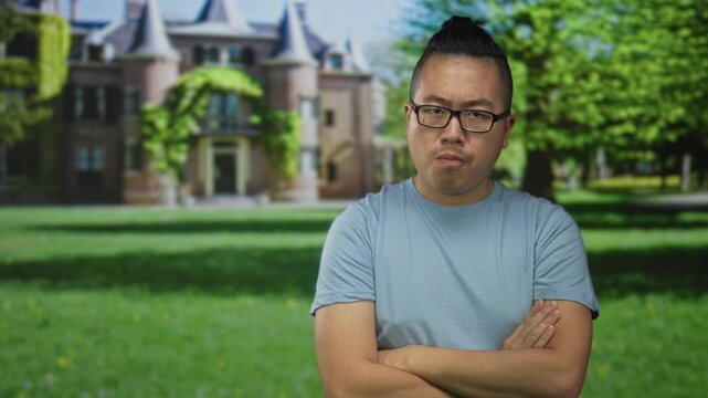 Young man with glasses and arms crossed in front of building and castle on green lawn; quiet introspection.