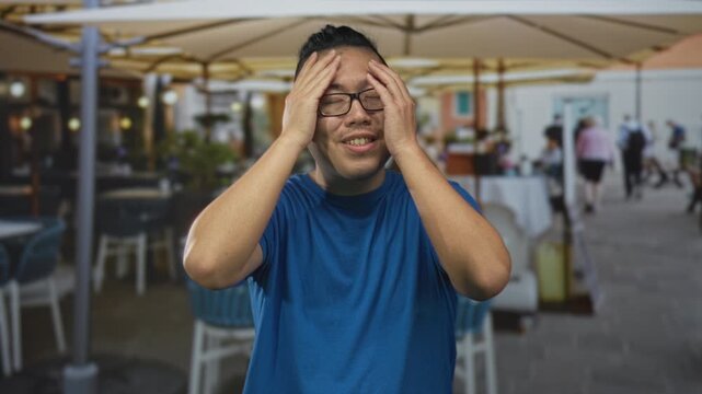 Man in blue shirt wearing glasses with hands on forehead on street terrace outside a restaurant under patio umbrellas, smiling and rubbing his brow; relief.