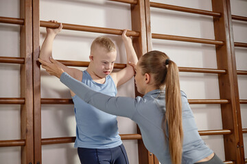 Caucasian boy child with Down syndrome exercising on wall bars with assistance from young Caucasian...