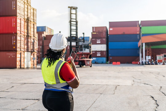 African female logistics manager in a hard hat using a walkie-talkie to coordinate container shipping operations at a busy industrial port terminal with reach stacker and cargo crates.