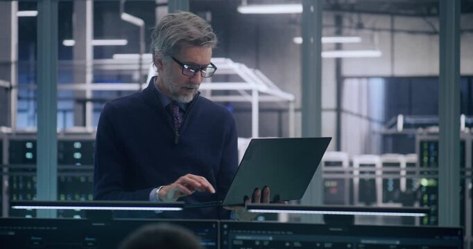 Focused Senior Manager in Formal Business Casual Wear Stands And Works, Uses Laptop to Analyze Data in Large, Mission Critical Server Room. Concept of Strategic Analysis and High Level IT Management.