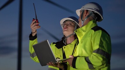 Team of professional male and female engineers in safety helmets and high-visibility jackets using a laptop and radio to inspect infrastructure or renewable energy site at dusk or night.
