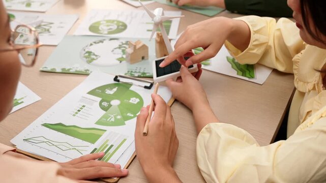 Business team focus windmill model, represented using clean energy. Coworker analyze green power graph choosing way to decrease carbon. Wooden block symbolized ecology house put on table. Delineation.
