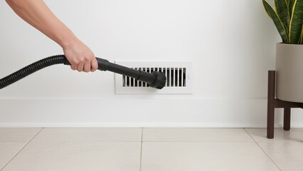 Close up of a person using a vacuum cleaner brush attachment to clean dust buildup from a low wall air vent