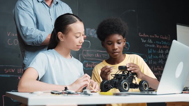 Caucasian teacher pointing at laptop screen while explain car model system at blackboard with coding or programming code written. Happy diverse student learning about robotic construction. Edification