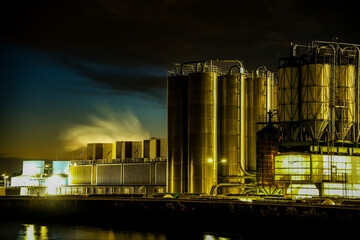 Gersthofen Industrial Park at night. Chemical production facility with large cylindrical silos, pipelines, and steaming emissions into the night sky. Reflection of artificial lighting in the Lech 