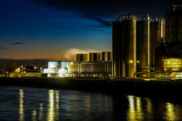 Gersthofen Industrial Park at night. Chemical production facility with large cylindrical silos, pipelines, and steaming emissions into the night sky. Reflection of artificial lighting in the Lech
