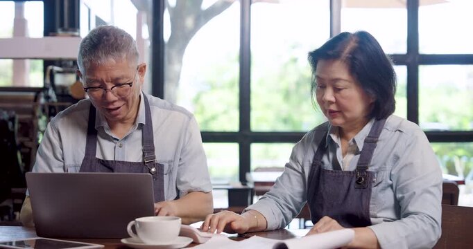 Asian elderly couple cafe owner in aprons working together on laptop writing notes at wooden table with coffee cups, teamwork management showing cooperation planning family entrepreneurs running small