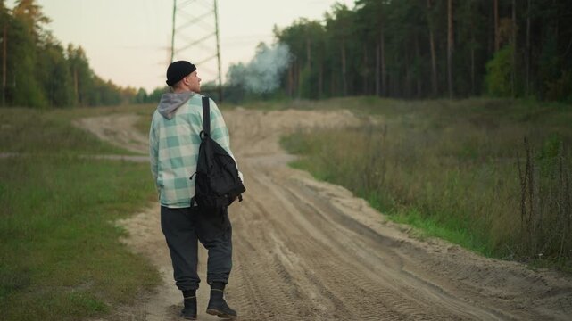 caucasian hiker turning on dirt road backpack slung over shoulder glancing back at path hesitant step toward crossroads tower silhouette grassy verge reflective pace rural exploration vibe