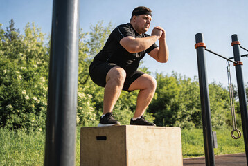 An overweight adult man wearing a black sports outfit performs a box jump in an outdoor gym under clear blue skies, surrounded by greenery and metal exercise equipment