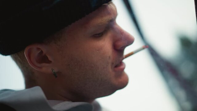white man traveler smoking in profile, beanie and earring, cigarette ember glowing against overcast sky closeup cinematic portrait shows stubble, weathered rail in background, introspective pause