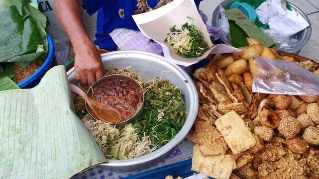 Footage of traditional Indonesian pecel salad with peanut sauce topping, packaged by a vendor at a traditional market, showing food preparation, simple wrapping, and authentic local market atmosphere.