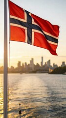 Norwegian flag proudly waving on a pole over tranquil waters at sunset, with a blurred city skyline in the background. Perfect for maritime-themed projects.