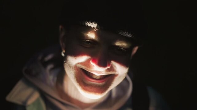 closeup flashlightlit caucasian face smiling, camp storyteller shifts roles teen camper, trail guide, urban explorer, latenight poet, prankster warm glow from handheld lantern illuminates hoodie,