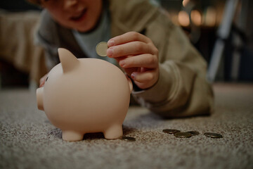 Caucasian child saving money by placing coin into piggy bank while lying on carpet, demonstrating financial awareness and early money management skills, coins scattered nearby