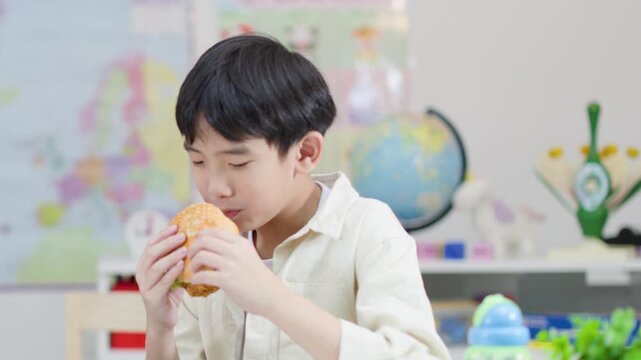 Young Asian Boy Enjoying a Large Chicken Burger in a Classroom