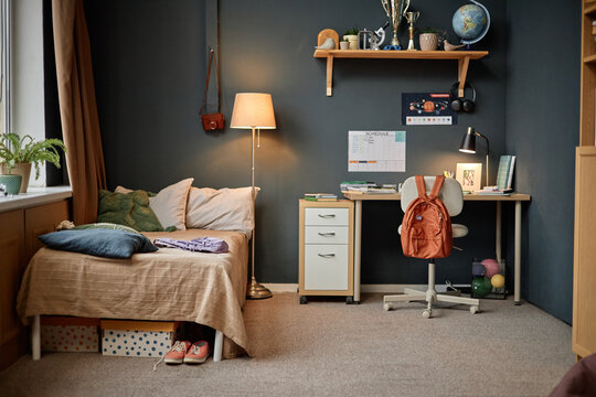 Cozy student bedroom with desk setup, books, backpack, and bed beside window, decorated with globe, plants, trophies, and a wall-mounted schedule 