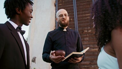 The priest gives the bride and groom wedding rings standing in front of the church at an outdoor...