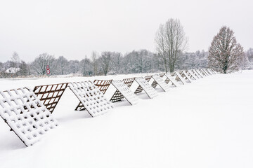 A winter landscape with snow-covered windbreak fence for snow stretching across a frozen field near the village, with a traffic sign visible in the background.