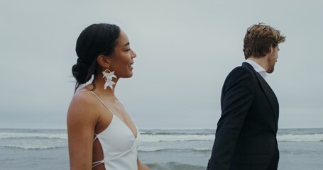 The bride and groom hold hands walking on the beach, the bride holds shoes in her hands