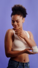 Young african american woman in a laconic top takes a cream from a jar and rubs it on her shoulder while posing against a monochrome studio background, vertical video