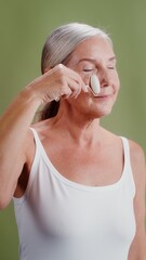 Elderly woman makes facial massage with a roller massager. Vertical video on monochrome studio background