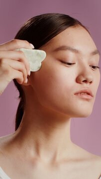 Young woman doing face massage with gua sha stone, posing in studio, vertical video