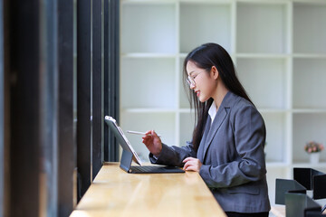 Professional businesswoman in eyeglasses working on a digital tablet with a stylus, standing at a counter near a window