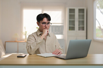 Young man wearing optical glasses using laptop and notebook, brainstorming a solution or strategy at his home office desk