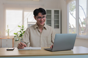 Young man smiling and wearing glasses, using a laptop and writing notes, sitting at a desk in a modern home office