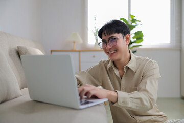 Young Asian man wearing eyeglasses smiling, using a laptop, and typing on the keyboard while sitting on the floor at home