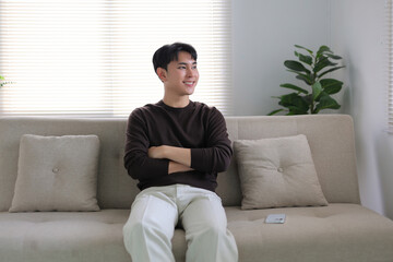 Young Asian man smiling contentedly, relaxing on a modern living room sofa in smart casual clothes, peaceful at home