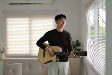 Young man smiling while playing acoustic guitar, enjoying music and relaxing in his comfortable living room