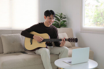 Happy young man practicing acoustic guitar, following an online tutorial on his laptop at home on the sofa