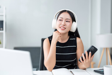 Asian woman wearing headphones and holding a smartphone while smiling and working remotely in a home office environment