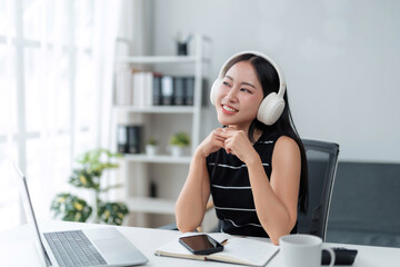 Young Asian woman wearing white headphones, smiling, listening to music or podcast while working in a bright modern office