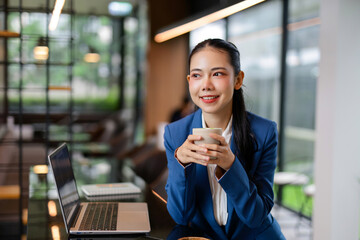 Asian businesswoman smiling, holding a cup of coffee. She is relaxing during a business break in a modern office or cafe