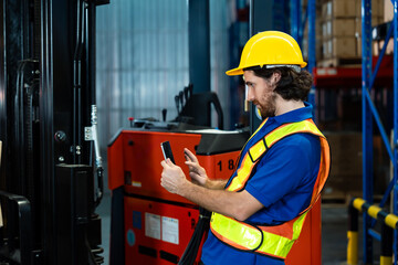 Male adult warehouse worker wearing yellow helmet and reflective vest using mobile device for inventory data entry near forklift inside modern distribution center, inside logistics warehouse. © Ekkasit A Siam