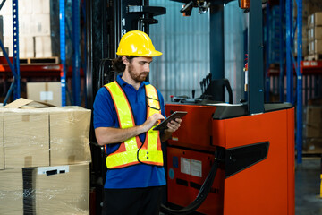 White male adult warehouse worker using digital tablet near forklift checking stock near stacked cardboard boxes wearing helmet and reflective vest in industrial storage space