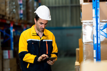 White adult male warehouse employee using tablet to check inventory inside storage space, focused on data while standing near packed boxes and tall shelves, inside logistics warehouse.
