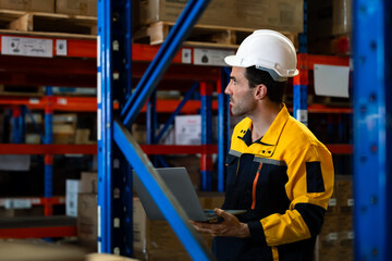 Caucasian male inventory staff wearing helmet using laptop to inspect stock inside shelf area of warehouse focusing on logistics data during routine check in distribution facility