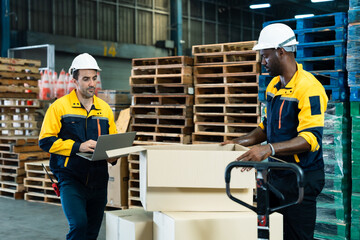 Asian adult male warehouse worker using laptop to manage inventory beside stacked cardboard boxes, standing in industrial space filled with pallets and shipping materials, inside logistics warehouse.