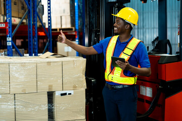African male warehouse worker wearing safety vest and helmet smiling while pointing and holding digital tablet beside stacked packages inside stockroom, inside logistics warehouse.