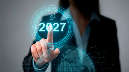 A businesswoman is touching a digital interface with the number 2027 and a sheet of paper, symbolizing the future.