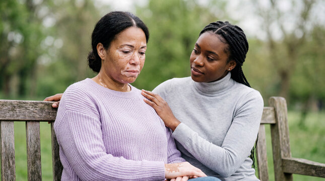 Caring young African woman offering support and comfort to a sad middle-aged friend with vitiligo on a park bench.