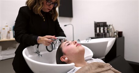 Professional hairstylist gently rinsing a relaxed female client's hair in a modern beauty salon wash basin before applying treatment, highlighting the pampering and hair care process
