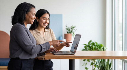 Two diverse businesswomen collaborating in a modern office, smiling and pointing at a laptop screen with a growth chart.