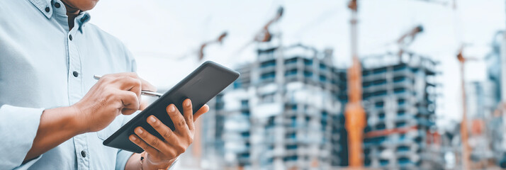 Construction supervisor uses tablet with cranes and building site blurred behind.